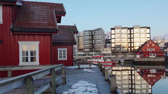 Footsteps echo along the harbor as morning light shines on red and gray buildings. The water is still, showing clear reflections of the structures. Boats are anchored nearby.