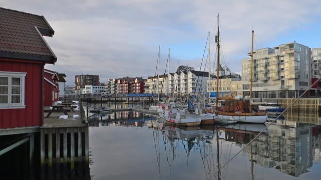 Footsteps echo along the harbor as morning light shines on red and gray buildings. The water is still, showing clear reflections of the structures. Boats are anchored nearby.