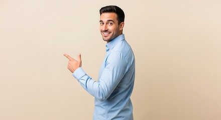Smiling young man in a light blue button-up shirt looking at camera and pointing his finger to the side at copy space, standing against a neutral beige background