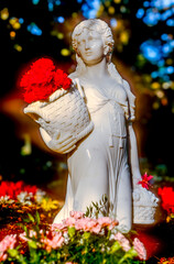 Analoges Foto einer Statuette eines M&auml;dchens mit mit Blumen gef&uuml;llten K&ouml;rben auf einem Friedhof im Licht der Sonne mit Weichzeichnereffekt