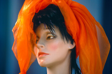 Kopfportrait einer Schaufensterpuppe mit orngenem Kopftuch und traurigem Blick vor unscharfem Hintergrund