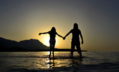 Romantic silhouette of a couple holding hands while standing in water during sunset. Love, freedom, summer vacation, and emotional connection concept.