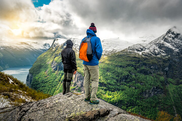 Two tourists are standing on the rock cliff over Geiranger fjord