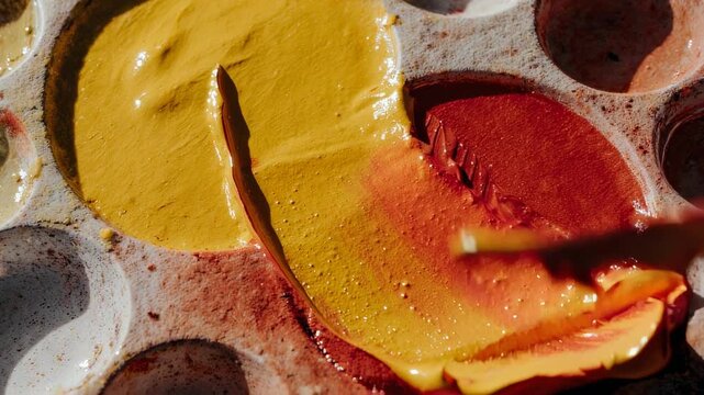 Artist Mixing Red and Yellow Paint On a Palette With a Metal Spatula During Daytime Natural Lighting