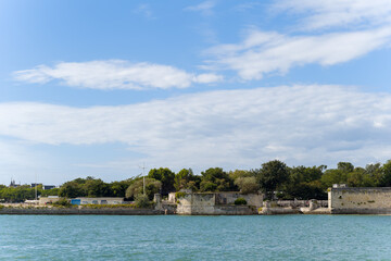 Fototapeta premium Historic stone walls and ramparts line the waterfront in La Rochelle, bordered by lush trees and set against a bright blue sky with scattered clouds. The tranquil scene captures the blend of heritage
