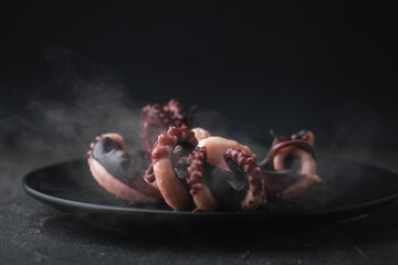 Hot prepared squid or boiled octopus with tentacles wiggling on a plate with steam coming out against black background. Seafood background for a restaurant menu © alexanderon