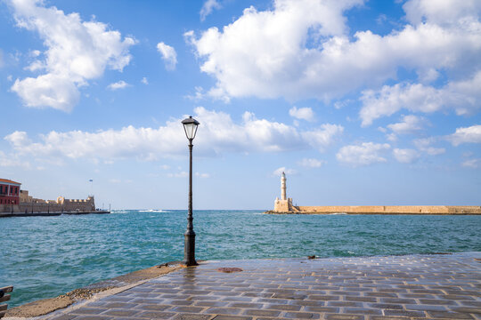 Historic lighthouse stands at the end of a stone pier in Chania's old Venetian harbor, with a classic lamppost and wet cobblestones in the foreground under a bright sky with scattered clouds.