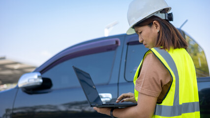 Worker in safety gear uses laptop while standing next to a parked truck at a construction site during the day. The area is active with work