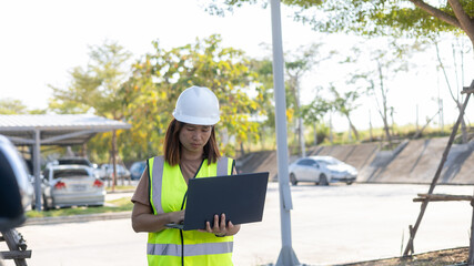 A woman in a hard hat and safety vest uses a laptop while standing in a parking area. Vehicles are parked in the background as she focuses on the screen. It is a sunny day