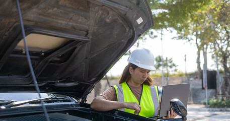 A woman wearing a safety vest and hard hat checks the status of a vehicle while using a laptop. She stands by a car with its hood open, surrounded by trees in daylight