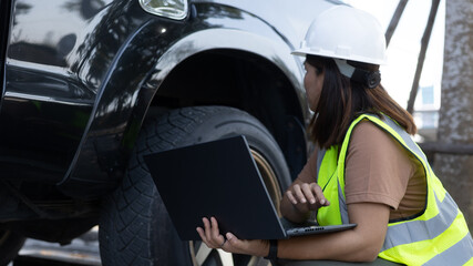 A worker wearing a hard hat and vest is checking a vehicle using a laptop. The black SUV is parked under a tree. The worker is focused on the task during the day