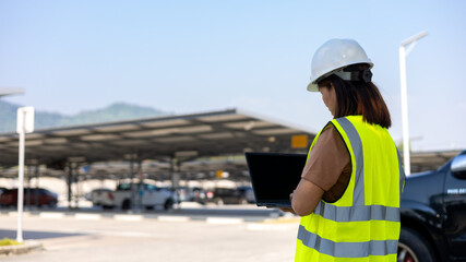 A worker wearing a safety vest and hard hat stands in a parking lot. The person is focused on using a laptop. Cars and mountains are seen in the background © Happy Photo