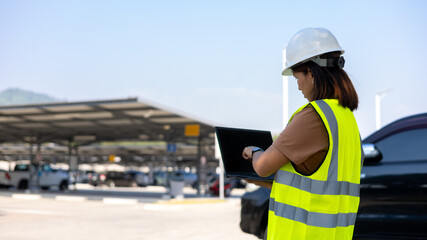 Worker stands in a parking lot wearing a safety vest and helmet. They are looking at a laptop and checking data. Cars are parked in the background under sunny skies