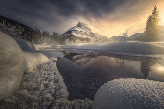 View of a mountain peak reflected in the calm waters surrounded by snow-covered banks and frosted trees under a warm sky, Banff, Alberta, Canada.