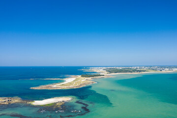 Fototapeta premium Drone image of a narrow sandy peninsula stretching into clear blue-green waters near Quiberon, with a small islet and the coastline visible under a vibrant sky. The scene is bright and expansive