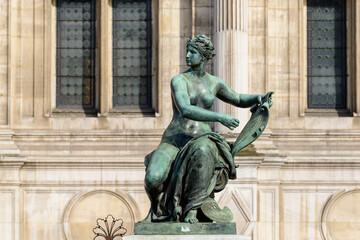 Fototapeta premium Patinated bronze sculpture of a seated woman holding a scroll, set against the historic stone facade and tall windows of Hotel de Ville in Paris, illuminated by soft natural light.