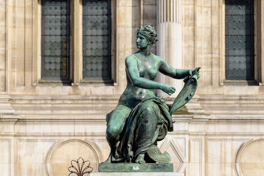 Patinated bronze sculpture of a seated woman holding a scroll, set against the historic stone facade and tall windows of Hotel de Ville in Paris, illuminated by soft natural light.