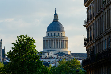 Naklejka premium Majestic dome of the Pantheon rises above rooftops, framed by historic Parisian architecture and lush green trees in soft evening light.