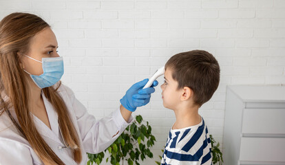 Female doctor in face mask measuring boy's temperature with infrared thermometer