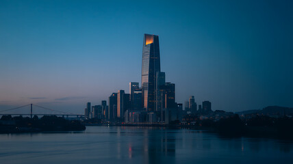 Obraz premium Dramatic Cityscape at Twilight: Modern Skyscrapers Reflecting in Calm River Waters Under a Deep Blue Sky at Dusk with Bridge in the Distance