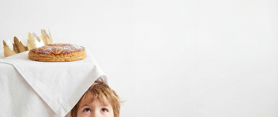 Playful child hiding under a table topped with a traditional Galette des Rois and golden crown. Minimalist Epiphany concept with a kid peeking at the French King Cake, featuring ample copy space.