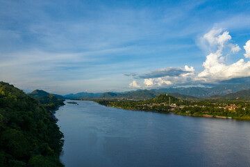 Fototapeta premium Expansive view of Luang Prabang city nestled between lush green hills and the wide Mekong river under a blue sky with dramatic clouds. The scene highlights tropical vegetation, distant mountain ranges