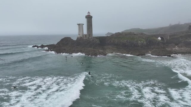 Aerial view of surfers riding waves near the historic Phare du Petit Minou lighthouse on a cloudy day, Bretagne, France.