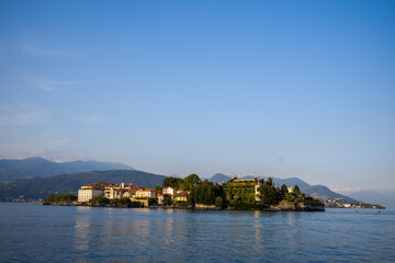 Fototapeta premium Warm evening sunlight bathes Isola Bella, highlighting the island's historic buildings and lush terraced gardens surrounded by the calm blue waters of Lake Maggiore. Distant alpine mountains and a