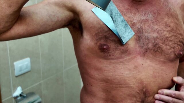 Humorous but painful shot of a mature man attempting a do-it-yourself epilation on his hairy chest using strong adhesive tape, showing an unconventional and unsuitable beauty treatment