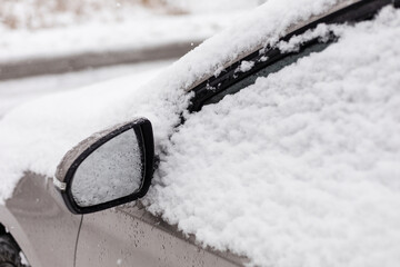 A parked car is covered with a thick layer of fresh snow. Large snowflakes fall gently, blanketing...