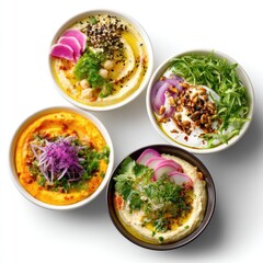 Assorted bowls of colorful hummus topped with fresh vegetables and herbs on a white background