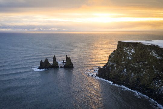 Aerial view of the stark, dark basalt sea stacks of Reynisdrangar rise from the turbulent waters beneath a golden, ethereal sky, Vik, Iceland.