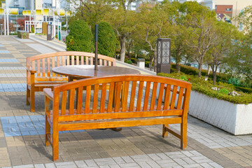 Set of wooden garden furniture with two benches and a round table on a paved city terrace. Bright outdoor setting with lush green trees and flower planters in a public recreational space.