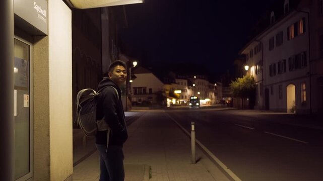 A person stands at a bus stop at night, with buildings and streetlights illuminating the dark surroundings Stock Video