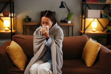 Sick Asian woman blowing nose with tissue wrapped in warm blanket