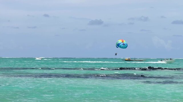 A person is parasailing high in the sky, enjoying the view over a beachfront area. The colorful parachute contrasts with the blue sky and white clouds.