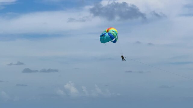A person is parasailing high in the sky, enjoying the view over a beachfront area. The colorful parachute contrasts with the blue sky and white clouds.