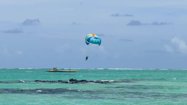 A person is parasailing high in the sky, enjoying the view over a beachfront area. The colorful parachute contrasts with the blue sky and white clouds.