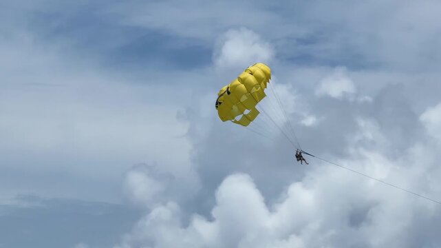 A person is parasailing high in the sky, enjoying the view over a beachfront area. The colorful parachute contrasts with the blue sky and white clouds.