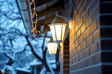 Warm glow of vintage lanterns illuminating a brick wall, adorned with colorful holiday lights, creating a cozy winter atmosphere in a snowy outdoor setting