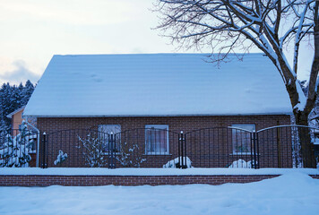 Snow-covered house with brick exterior and white roof, surrounded by a snowy landscape and bare trees, creating a serene winter atmosphere with peaceful ambiance