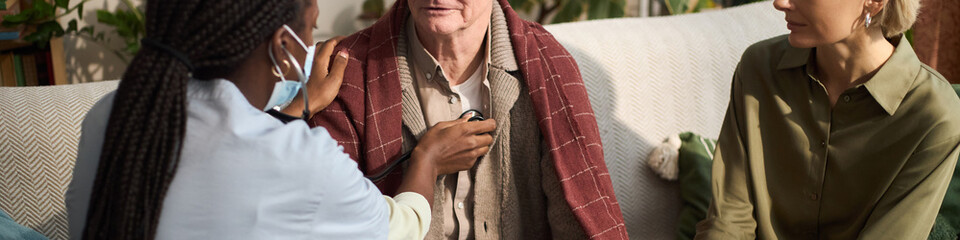 Header of Senior Caucasian man sitting on sofa being examined by Black female healthcare worker...