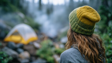 Person in vibrant green beanie standing on forest ground, gazing at a tent ahead, mist swirling at ground level, tranquil outdoor exploration atmosphere