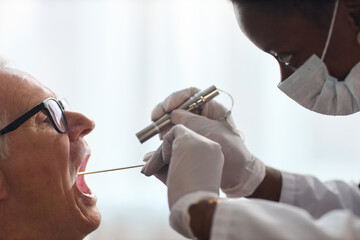 Senior Caucasian man sitting with mouth open, while Black woman medical professional wearing mask and gloves, examining throat using tongue depressor and flashlight in clinical setting