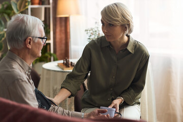 Middle aged Caucasian woman comforting senior Caucasian man while sitting together indoors, woman holding man's arm and man holding cup, both engaged in serious conversation