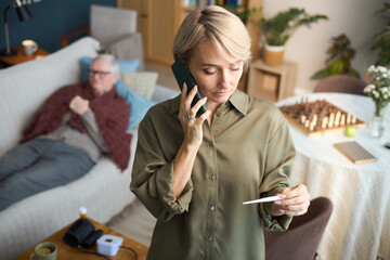 Caucasian middle aged woman talking on smartphone while holding thermometer, senior Caucasian man...