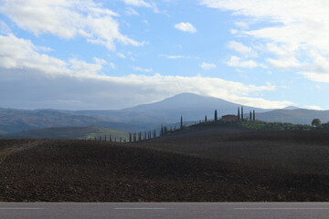 A stunning Tuscan landscape showcasing rolling hills, lined with tall cypress trees, under a bright blue sky. The peaceful countryside and distant mountains create a serene and beautiful scene.