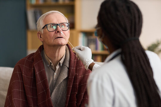 Caucasian senior man sitting while Black woman healthcare worker performing nasal swab test, both indoors, man wearing glasses and looking upward, medical examination in progress