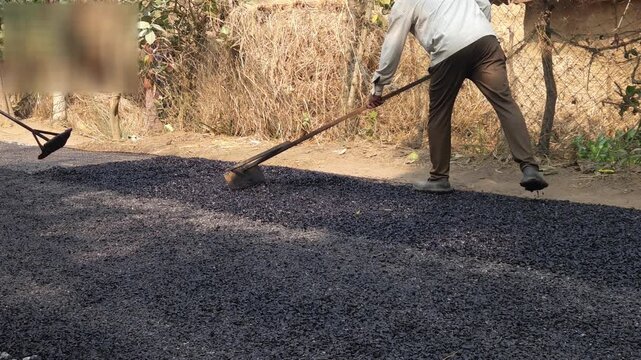 Construction worker manually spreads hot asphalt on a rural road using traditional tools, highlighting labor-intensive road construction practices in India.