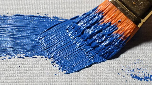 Close up of an orange handled paintbrush with blue paint creating a thick stroke on a textured white canvas surface with detailed bristle marks visible and a slightly blurred background highlighting